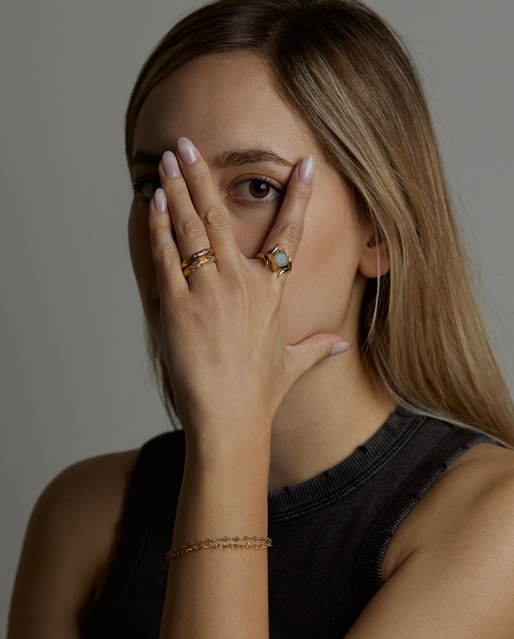 A portrait of a woman model covering her face with her hand. On her hand she is wearing the light and shadow rings with topaz natural gemstones and the ascension ring with natural aquamarine gemstone. On her wrist she wears the hearts double chain labradorite bracelet. All jewelry are made of silver and gold vermeil. Demi-fine jewelry. Made in Athens, Greece, Europe,EU.