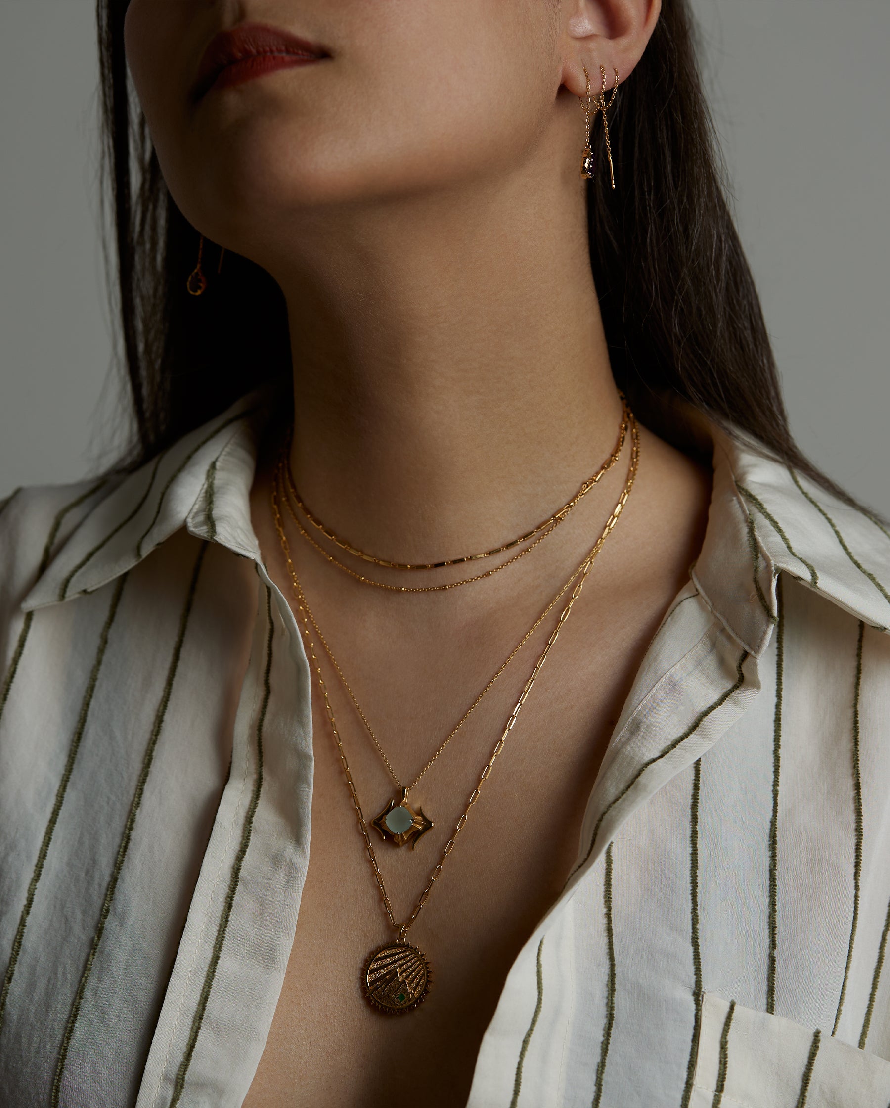 A closeup portrait of a woman model's neck wearing the ray double necklace, the ascension necklace with the aquamarine natural gemstone and the terra element necklace with an emerald natural gemstone. On her ear she wears the soul earring with natural amethyst gemstone. All jewelry are made of silver and gold vermeil. Demi-fine jewelry. Made in Athens, Greece, Europe,EU.