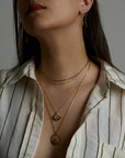 A closeup portrait of a woman model's neck wearing the ray double necklace, the ascension necklace with the aquamarine natural gemstone and the terra element necklace with an emerald natural gemstone. On her ear she wears the soul earring with natural amethyst gemstone. All jewelry are made of silver and gold vermeil. Demi-fine jewelry. Made in Athens, Greece, Europe,EU.