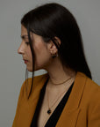 A portrait of a woman model in a mustard suit that on her ear is wearing three earrings. The essence big earrings with natural onyx gemstones and two essence hoop earrings with a natural sodalite gemstone and a natural rose quartz gemstone. On her neck she wears the cosmos necklace with a natural black onyx gemstone and the ray double chain necklace. All jewelry are made of silver and gold vermeil. Demi-fine jewelry. Made in Athens, Greece, Europe,EU.