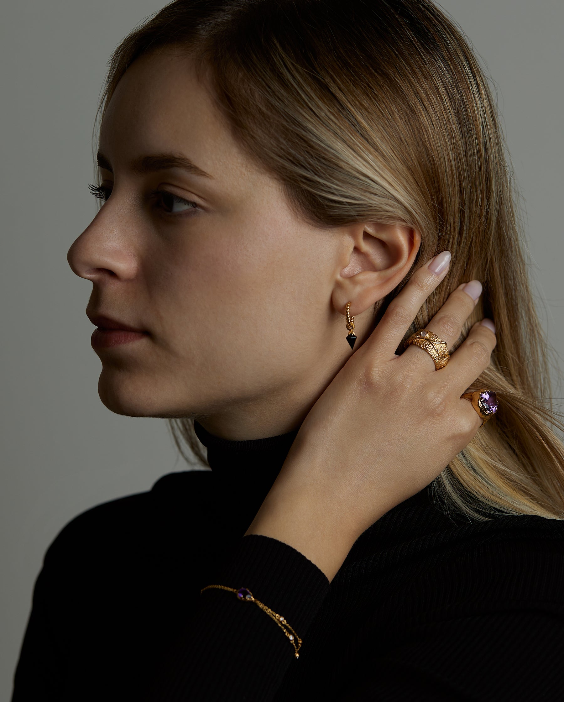 A portrait of a woman model brushing her hair with her hand. She is wearing many jewelry. On her wrist she has the soul bracelet with natural amethyst gemstone. On her finger she wears two rings, the soul ring with natural amethyst gemstone and the messenger ring with a natural chalcedony purple gemstone. On her ear she wears the essence hoop earring with natural onyx gemstone. All jewelry are made of silver and gold vermeil. Demi-fine jewelry. Made in Athens, Greece, Europe,EU.