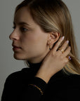 A portrait of a woman model brushing her hair with her hand. She is wearing many jewelry. On her wrist she has the soul bracelet with natural amethyst gemstone. On her finger she wears two rings, the soul ring with natural amethyst gemstone and the messenger ring with a natural chalcedony purple gemstone. On her ear she wears the essence hoop earring with natural onyx gemstone. All jewelry are made of silver and gold vermeil. Demi-fine jewelry. Made in Athens, Greece, Europe,EU.