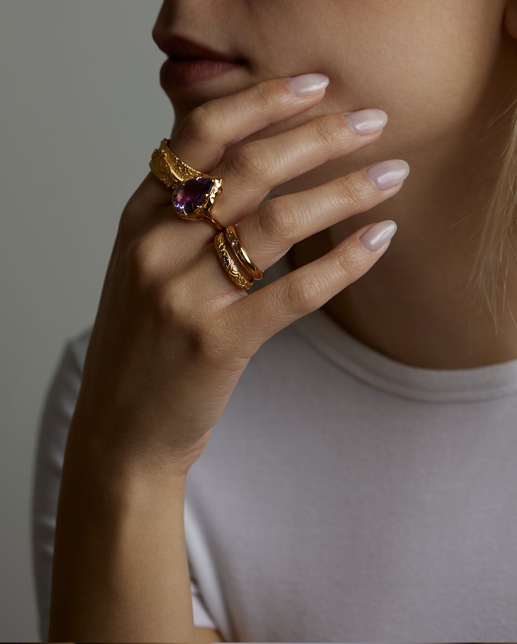 A portrait of a woman model holding her face with her hand. On the fingers she wears many rings, the soul ring with natural amethyst gemstone and the messenger ring with a natural chalcedony purple gemstone and the light and shadow rings with London blue topaz natural gemstones. All jewelry are made of silver and gold vermeil. Demi-fine jewelry. Made in Athens, Greece, Europe,EU.