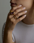 A portrait of a woman model holding her face with her hand. On the fingers she wears many rings, the soul ring with natural amethyst gemstone and the messenger ring with a natural chalcedony purple gemstone and the light and shadow rings with London blue topaz natural gemstones. All jewelry are made of silver and gold vermeil. Demi-fine jewelry. Made in Athens, Greece, Europe,EU.