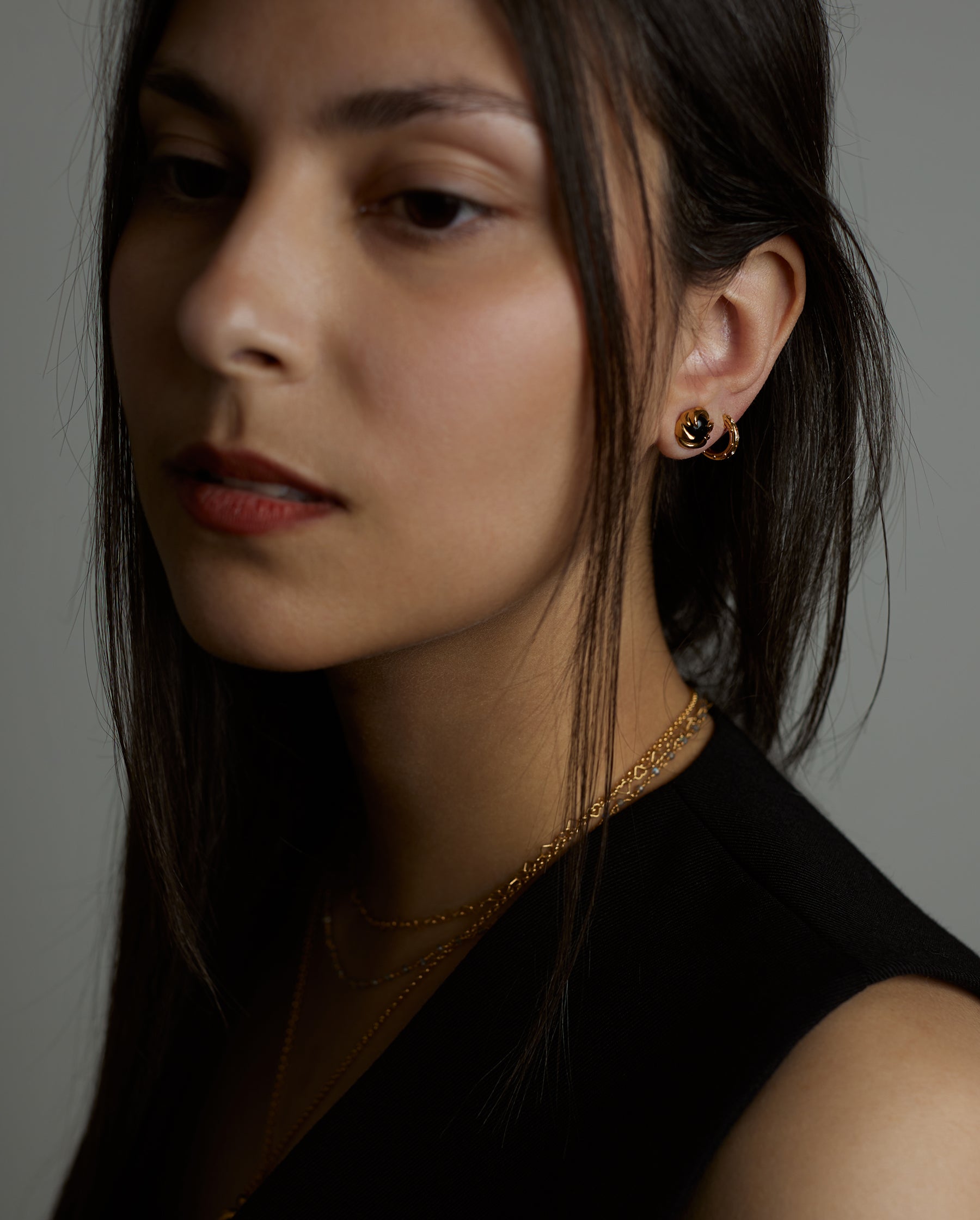 A portrait of a woman model that on her ear is wearing two earrings. The luck hoop earrings with natural topaz gemstones and the cosmos earrings with natural black onyx gemstones. All jewelry are made of silver and gold vermeil. Demi-fine jewelry. Made in Athens, Greece, Europe,EU.