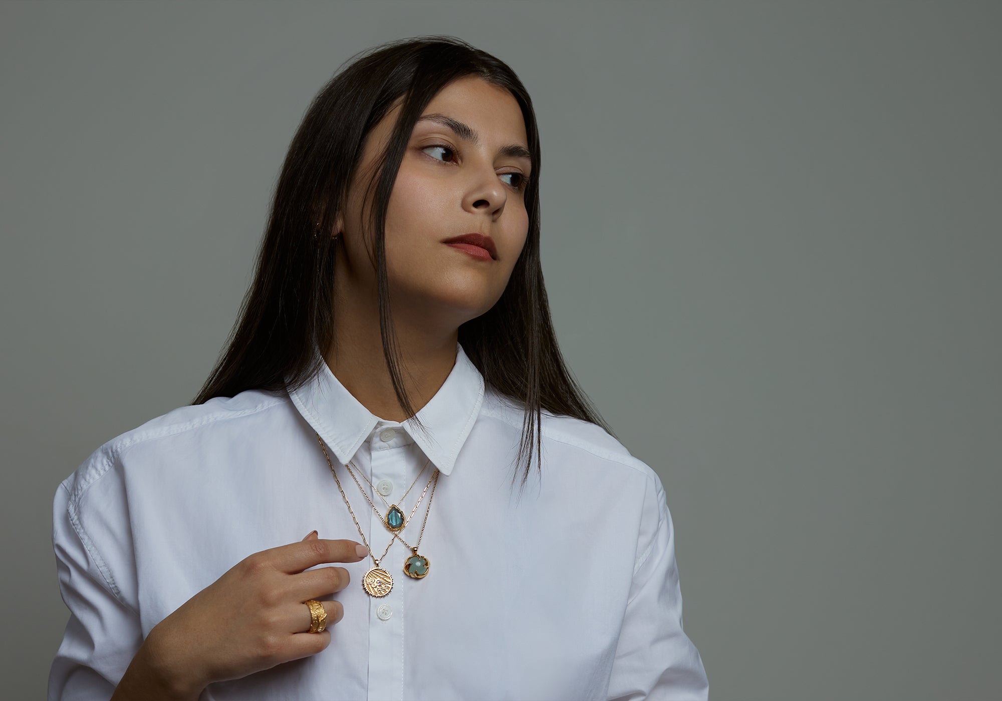 A portrait of a woman model with white shirt, wearing 3 necklaces. The soul necklace with a blue labradorite natural gemstone, the cosmos necklace with an aquamarine natural gemstone and the aer element necklace with an amethyst natural gemstone. On her ring she is wearing the messenger ring with a purple chalcedony natural gemstone. All jewelry are made of silver and gold vermeil. Demi-fine jewelry. Made in Athens, Greece, Europe,EU.