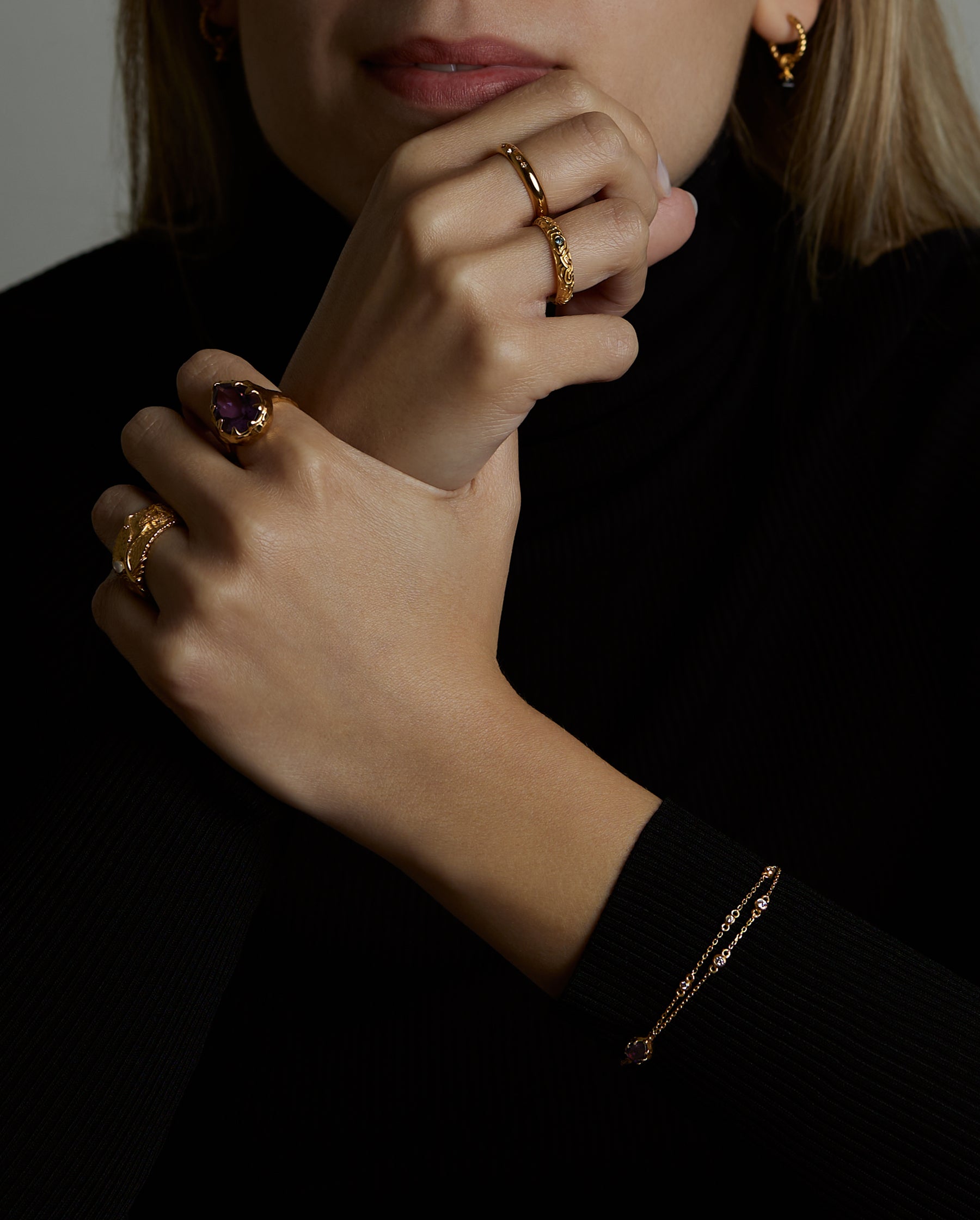 A portrait of a woman model holding her hands. She is wearing many jewelry. On her wrist she has the soul bracelet with natural amethyst gemstone. On the fingers of one hand she wears two rings, the soul ring with natural amethyst gemstone and the messenger ring with a natural chalcedony purple gemstone. On the other hand she wears the light and shadow rings with London blue topaz natural gemstones. On her ear she wears the essence hoop earring with natural onyx gemstone.