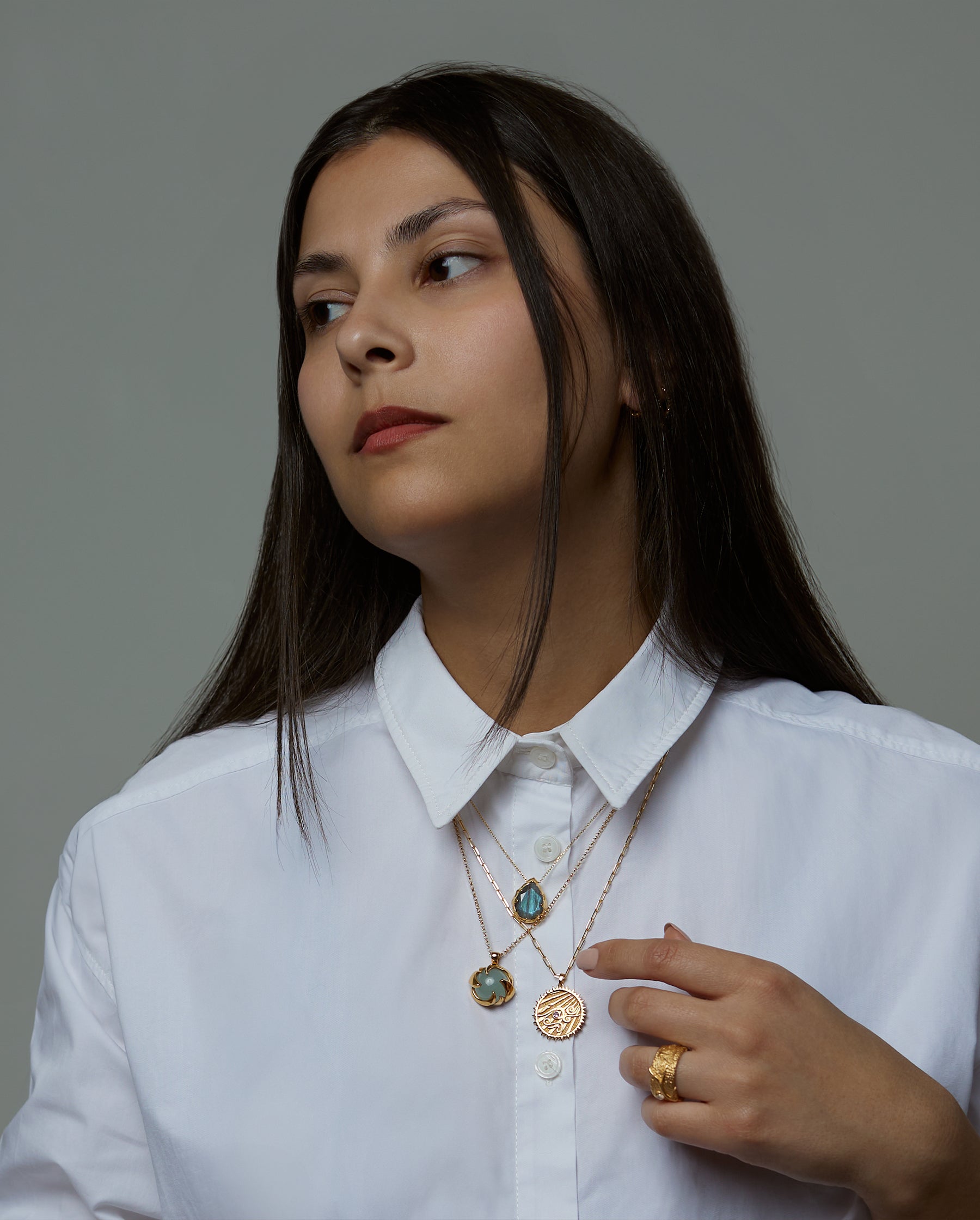 A portrait of a woman model wearing 3 necklaces. The soul necklace with a blue labradorite natural gemstone, the cosmos necklace with an aquamarine natural gemstone and the aer element necklace with an amethyst natural gemstone. On her ring she is wearing the messenger ring with a purple chalcedony natural gemstone. All jewelry are made of silver and gold vermeil. Demi-fine jewelry. Made in Athens, Greece, Europe,EU.