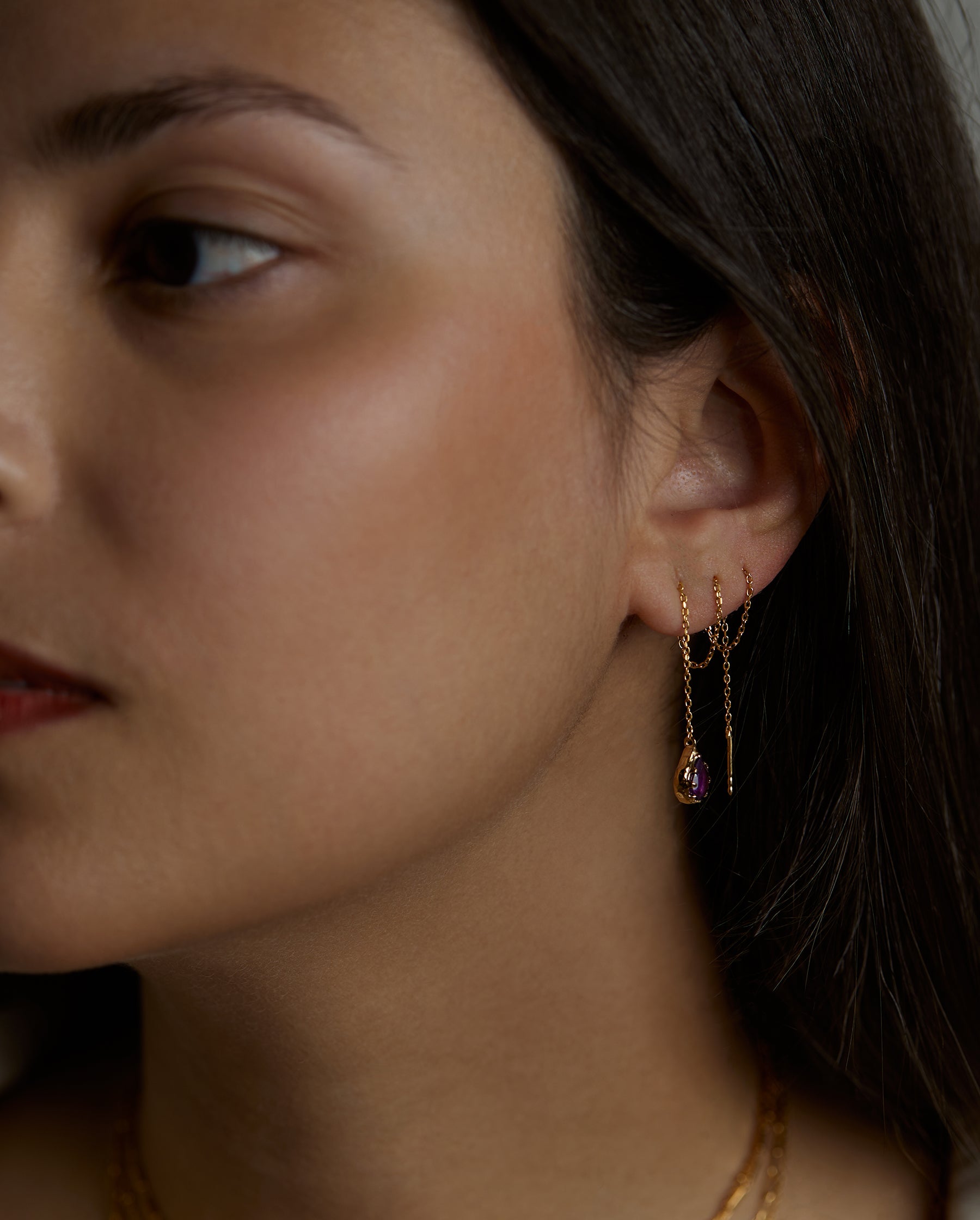 A portrait of a woman model that on her ear is wearing the soul earrings with natural amethyst purple gemstones. This earring is a long thread chain that she thread in three piercing holes of her ear. All jewelry are made of silver and gold vermeil. Demi-fine jewelry. Made in Athens, Greece, Europe,EU.