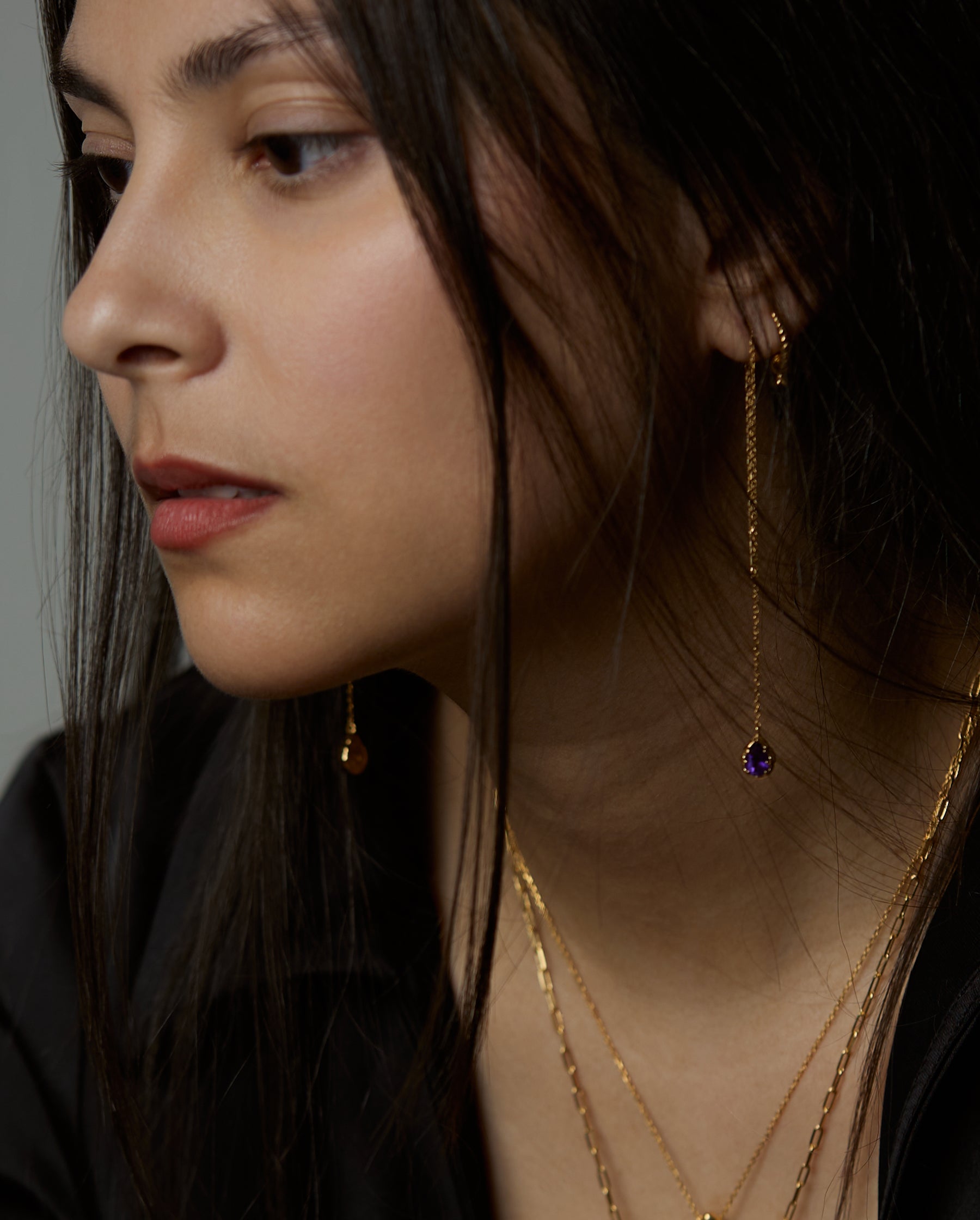 A portrait of a woman model that on her ear is wearing the soul earrings with natural amethyst purple gemstones. All jewelry are made of silver and gold vermeil. Demi-fine jewelry. Made in Athens, Greece, Europe,EU.