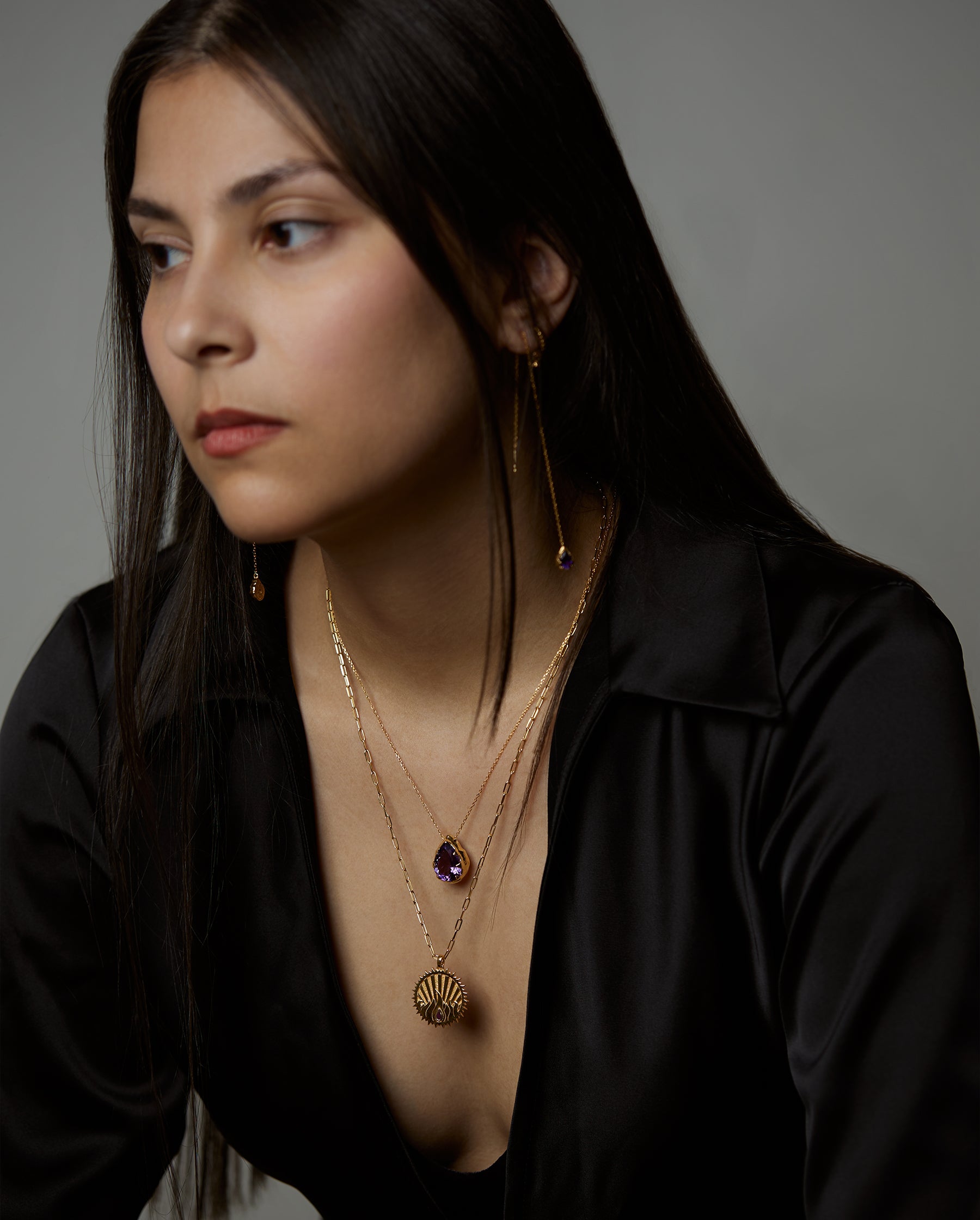 A portrait of a woman model wearing the soul necklace and soul earrings where both pieces have amethyst natural gemstones and the ignis element necklace with a ruby natural gemstone. All jewelry are made of silver and gold vermeil. Demi-fine jewelry. Made in Athens, Greece, Europe,EU.