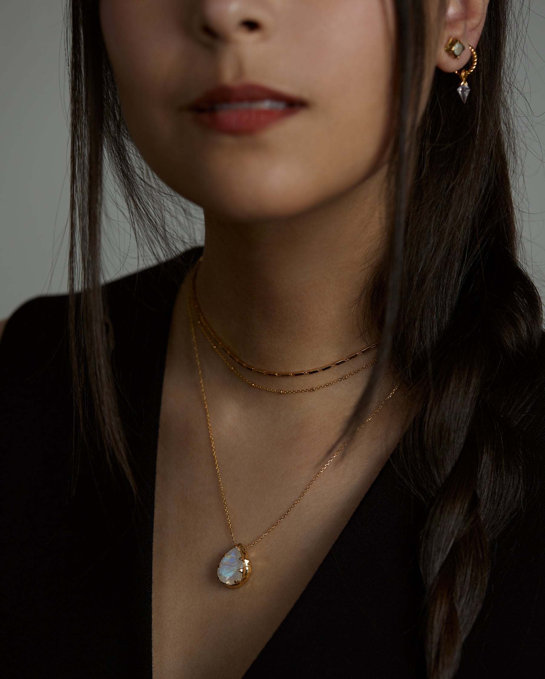 A closeup portrait of a woman model wearing the ray double necklace, the soul necklace with a moonstone natural gemstone, the ascension earring with aquamarine natural gemstone and the essence hoop earrings with rose quartz natural gemstones. All jewelry are made of silver and gold vermeil. Demi-fine jewelry. Made in Athens, Greece, Europe,EU.