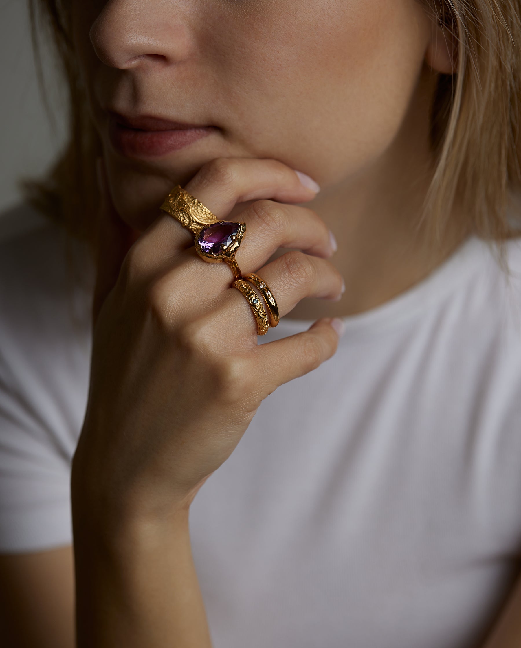 A portrait of a woman model holding her face with her hand. On the fingers she wears four different rings. The soul ring with natural amethyst gemstone and the messenger ring with a natural chalcedony purple gemstone and the light and shadow rings with London blue topaz natural gemstones. All jewelry are made of silver and gold vermeil. Demi-fine jewelry. Made in Athens, Greece, Europe,EU.