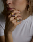 A portrait of a woman model holding her face with her hand. On the fingers she wears four different rings. The soul ring with natural amethyst gemstone and the messenger ring with a natural chalcedony purple gemstone and the light and shadow rings with London blue topaz natural gemstones. All jewelry are made of silver and gold vermeil. Demi-fine jewelry. Made in Athens, Greece, Europe,EU.